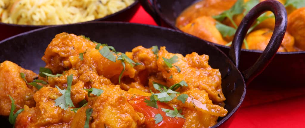 A close-up of chicken curry garnished with fresh coriander in a black bowl, with bowls of yellow rice and another curry visible in the background.