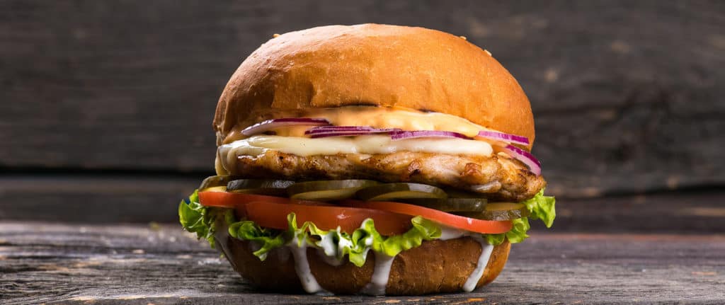 A grilled chicken sandwich with melted cheese, red onion, lettuce, tomato, gherkins, and mayonnaise in a toasted bap, set against a rustic wooden background.