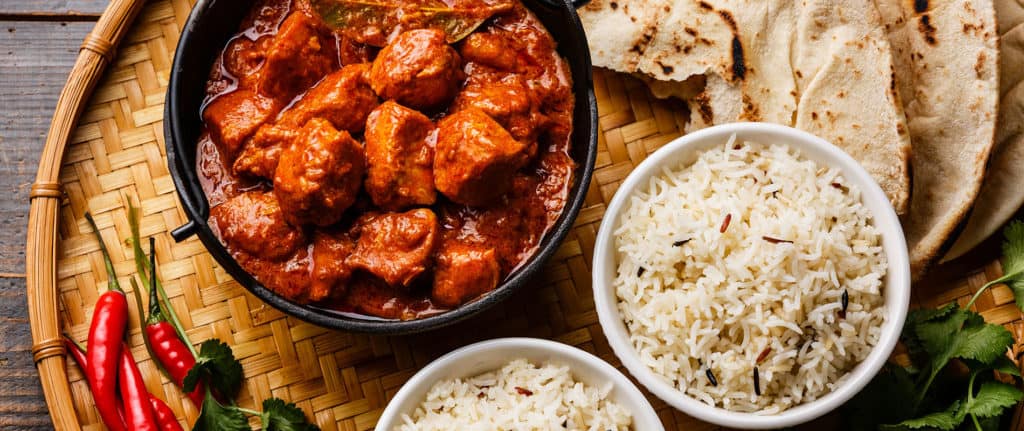 A bowl of chicken curry in rich red sauce sits on a woven tray next to two bowls of white rice, naan bread, red chilli peppers, and fresh coriander.