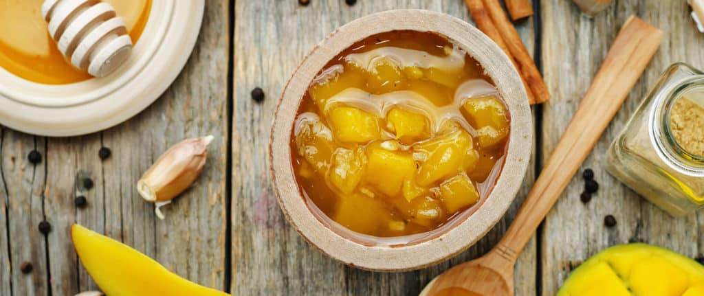 A wooden bowl filled with chunky mango chutney sits on a rustic wooden table, surrounded by spices, honey, a garlic clove, and fresh mango slices.