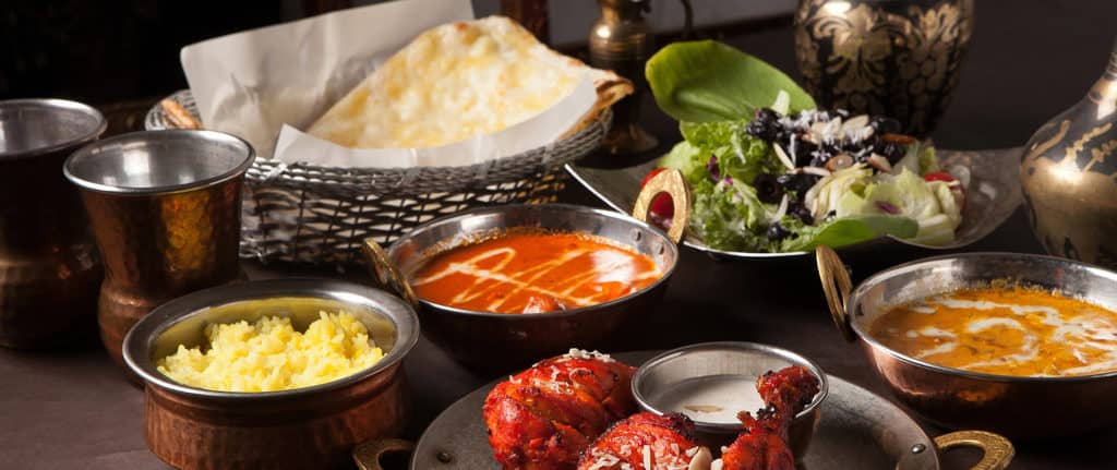 A table set with Indian cuisine, including naan bread, salad, bowls of yellow rice, creamy curries, and tandoori chicken, all served in traditional metal dishes and cups.