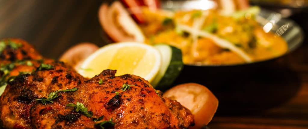 Close-up of grilled, seasoned chicken garnished with herbs and served with sliced lemon, onion, and cucumber, with a dish of curry in the blurred background.